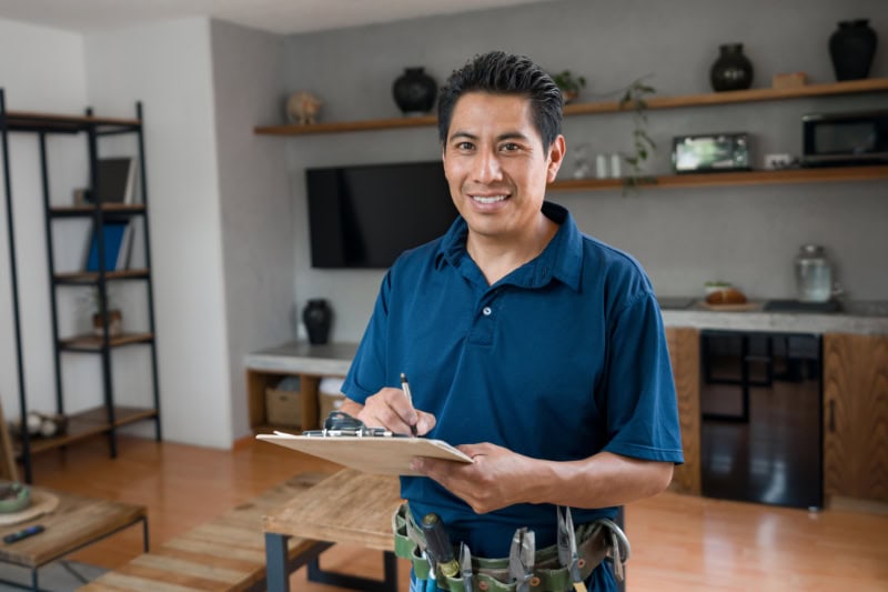 What Is a Plumbing Inspection? Happy electrician working at a house and looking at the camera smiling while writing on a clipboard - home improvement concepts.